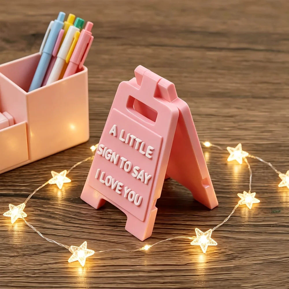 A pink 3D lettering sign shaped like an arrow with the text A LITTLE SIGN TO SAY I LOVE YOU resting on a wooden surface.