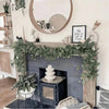 An artificial eucalyptus garland with white flowers draped across a fireplace mantel and surrounding decor.