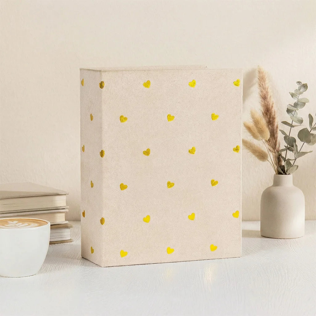 A beige photo album with gold foil hearts rests on a white surface next to a cup and dried floral arrangement.