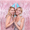 Two young women smiling and clapping in front of a pink metallic foil tassel backdrop set