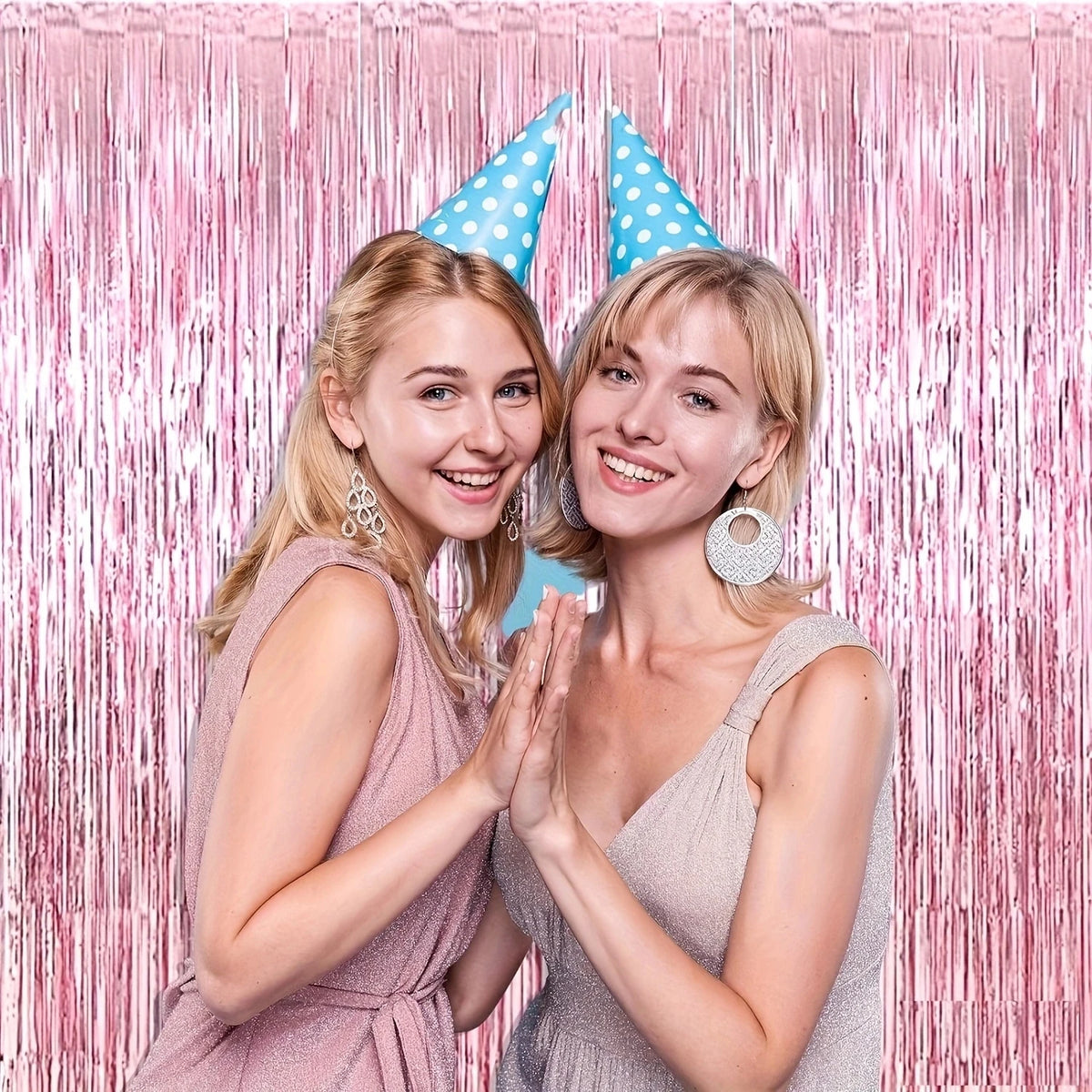 Two young women smiling and clapping in front of a pink metallic foil tassel backdrop set