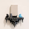 A dark, skull-shaped floating wall shelf displaying a light gray rectangular object and a blue glass sculpture.