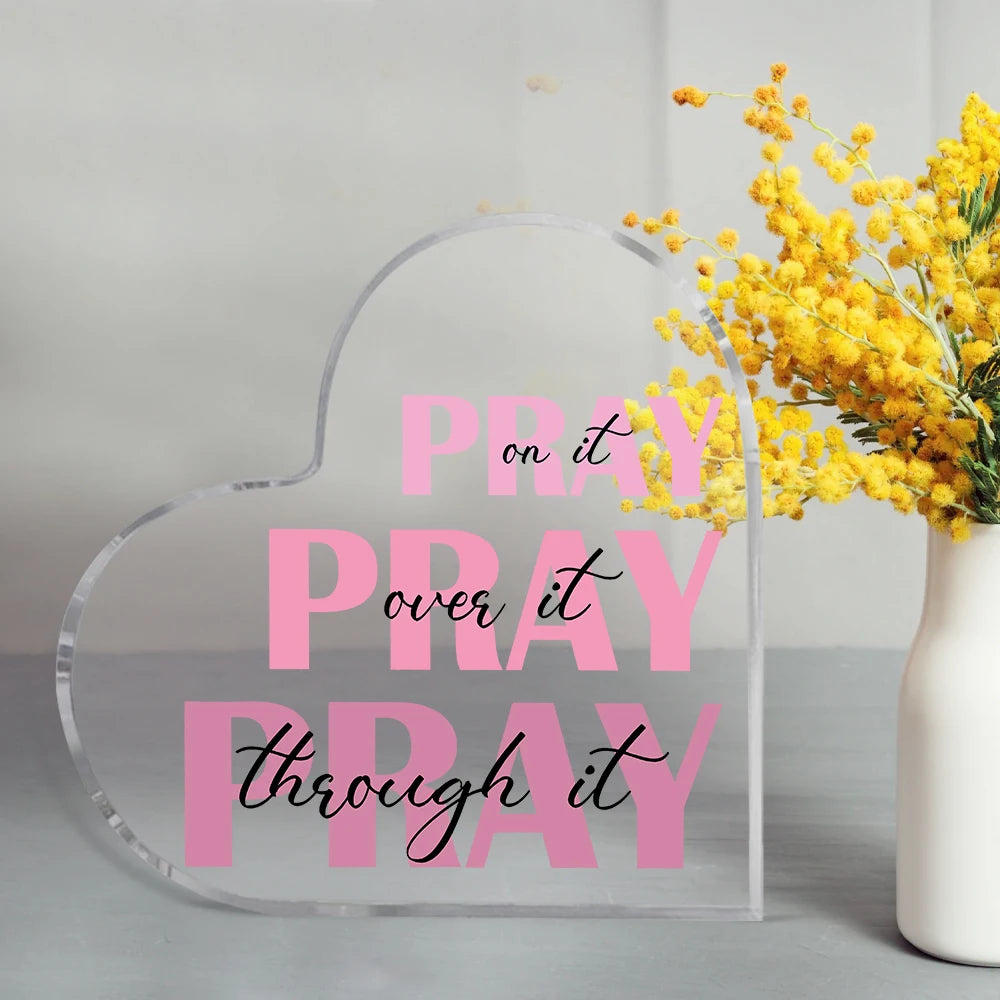 A clear acrylic heart-shaped sign with pink text that reads Pray it over it pray through it next to yellow flowers