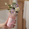 A handmade floral bouquet featuring purple and white flowers wrapped in pink paper and tied with pink ribbon.