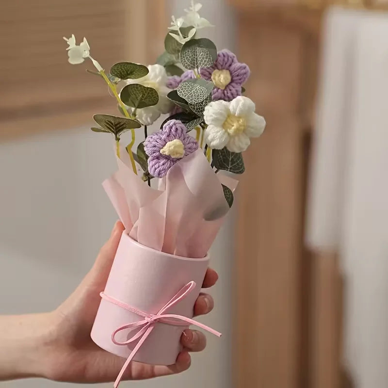A handmade floral bouquet featuring purple and white flowers wrapped in pink paper and tied with pink ribbon.