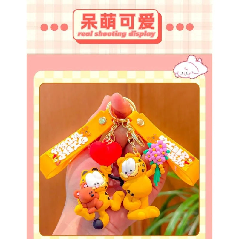 A hand holds a yellow Garfield cartoon keychain pendant with a red heart and pink flowers.