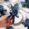 A hand holds a dusty blue silk rose boutonniere featuring blue thistle and lavender sprigs.