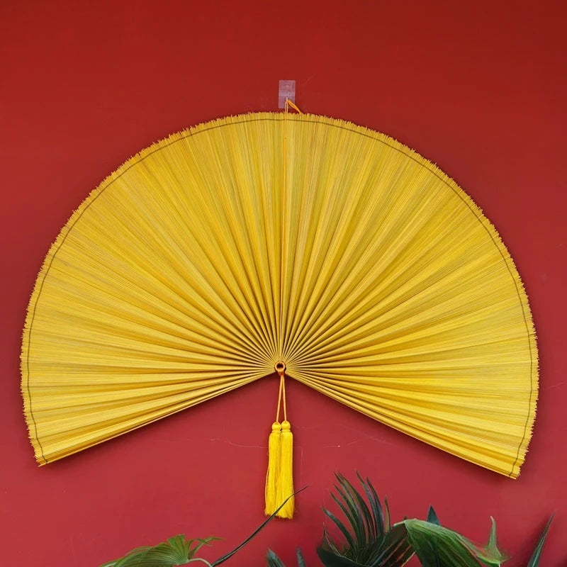 A bright yellow handwoven bamboo wall fan decor with a tassel displayed against a red background.