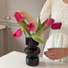 A hand holds a dark glass flower vase filled with vibrant pink and magenta tulips on a white countertop.
