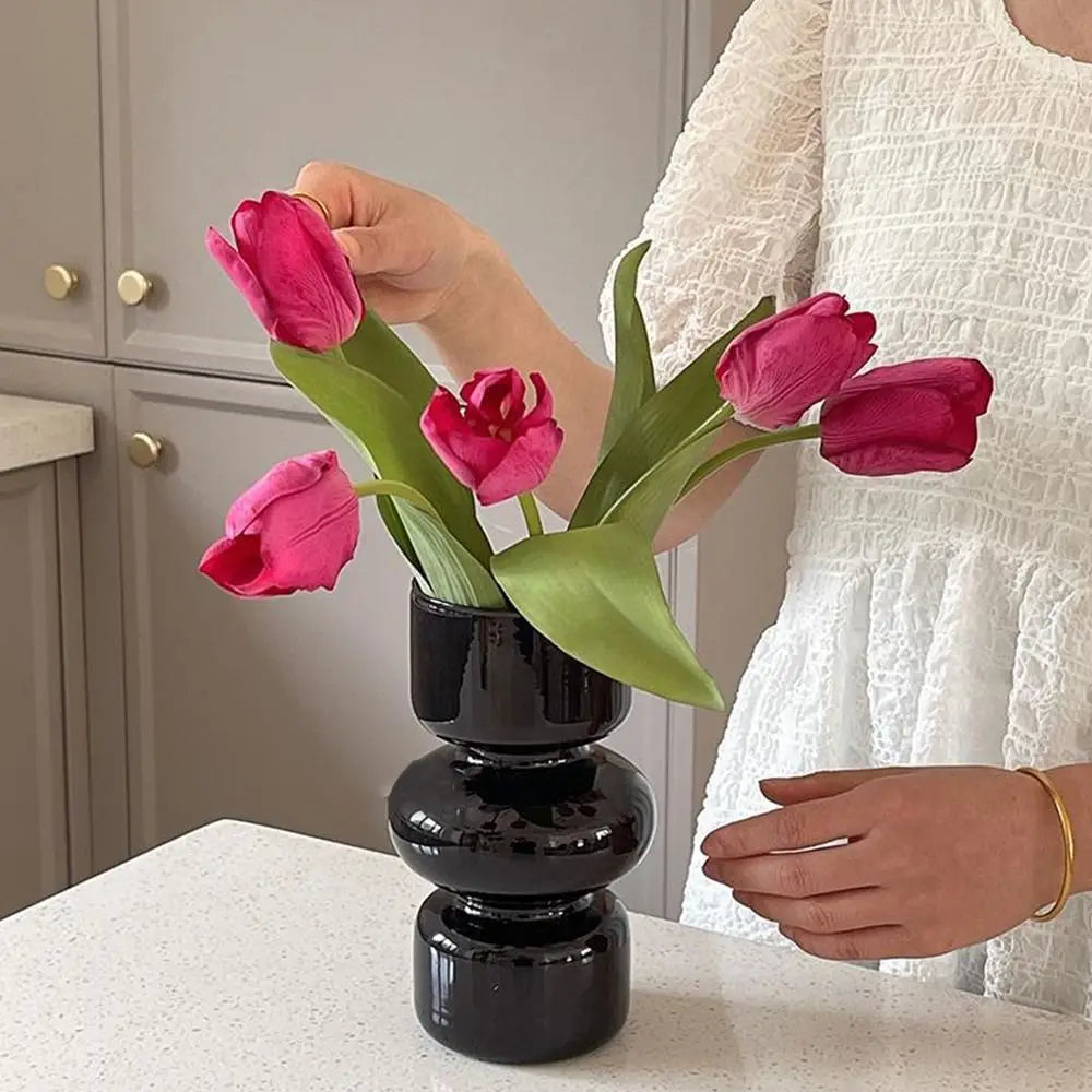 A hand holds a dark glass flower vase filled with vibrant pink and magenta tulips on a white countertop.
