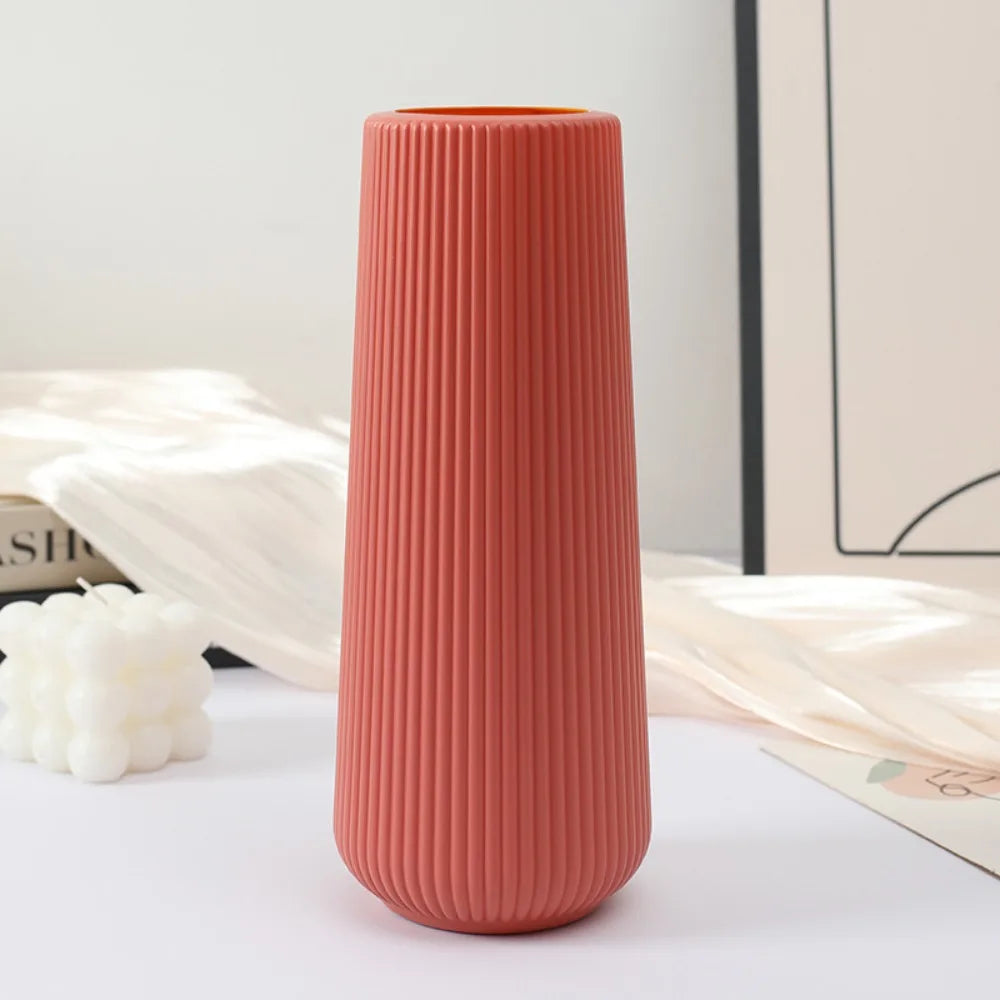 A tall, ribbed coral pink plastic vase with an orange rim is displayed on a white surface next to dried light beige plant mat