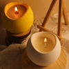 A yellow silicone arc candle jar with a lit candle sits on a wooden base next to a white ceramic candle jar.