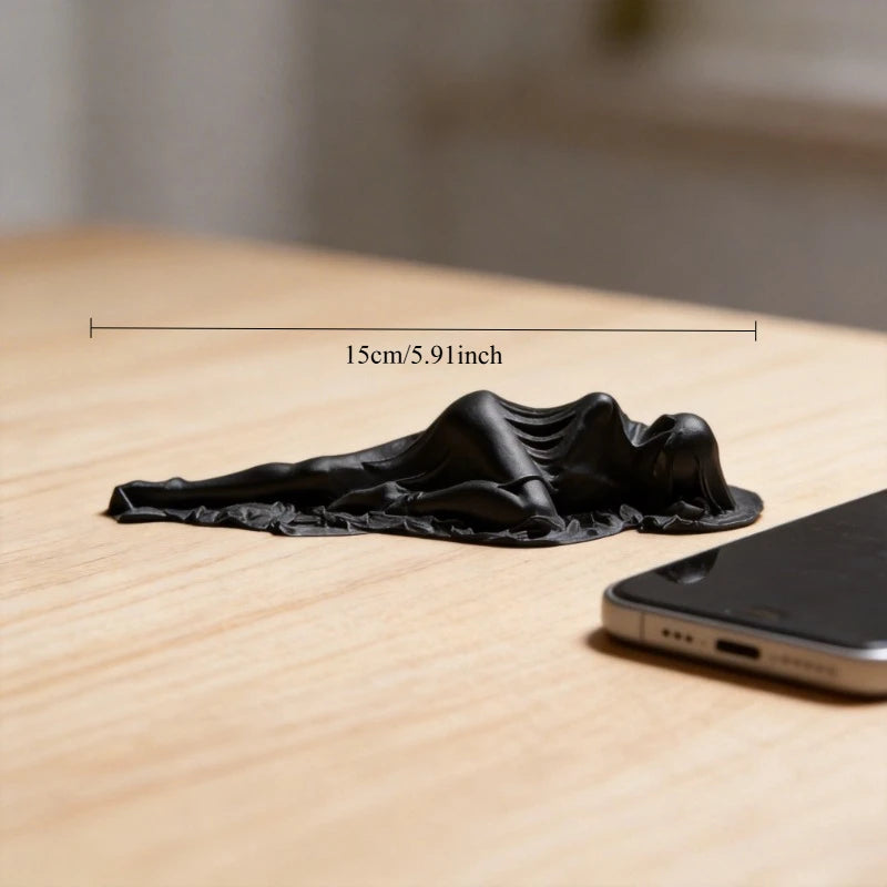 A black, draped reclining female statue sculpture is shown on a wooden desk next to a smartphone.