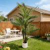 An artificial triple trunk palm tree in a white pot standing in a green lawn in front of a wooden fence and house.