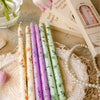 A collection of decorative printed long stick candles in various shades of purple and green are displayed on a lace doily.