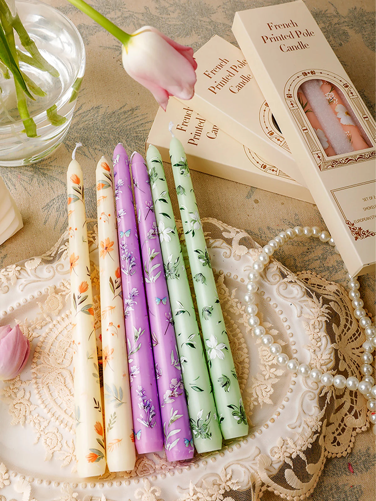 A collection of decorative printed long stick candles in various shades of purple and green are displayed on a lace doily.
