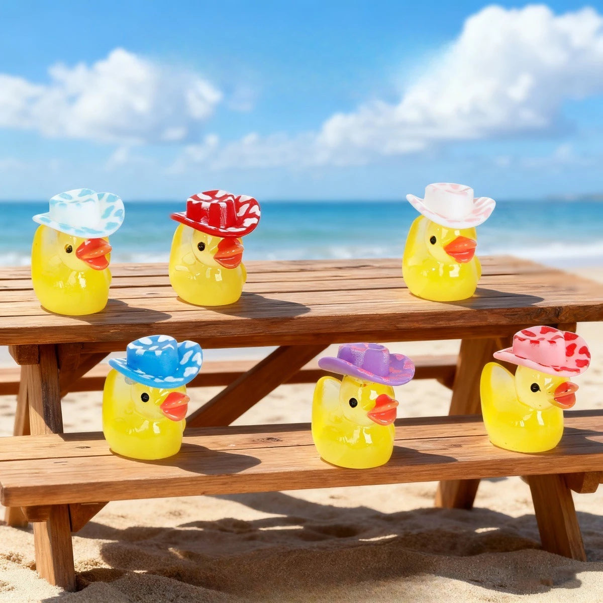 Three yellow resin duck figurines wearing colorful hats sitting on a wooden table near the ocean.