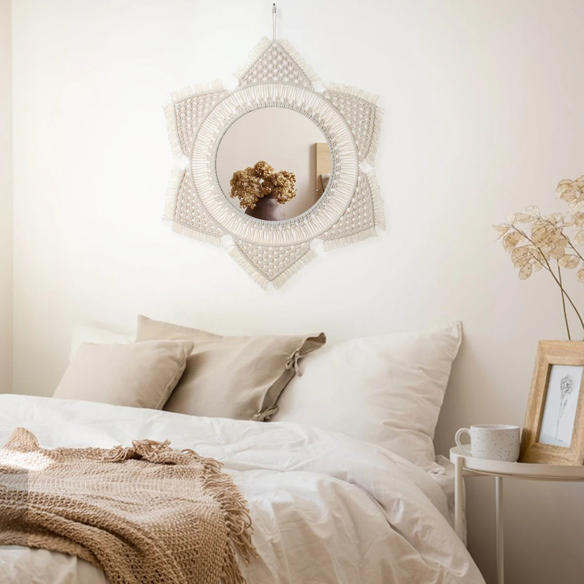 A round macrame wall mirror with a geometric frame hangs on a white wall above a bed.