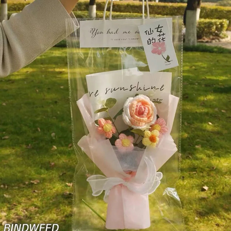 A handmade crochet flower bouquet featuring pink and yellow roses and filler flowers, presented in a clear plastic bag.