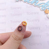 A hand holds a small, round resin charm shaped like a donut with cheese on it against a white background.