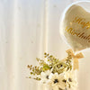A clear bubble balloon shaped like a heart with gold lettering that reads Happy Birthday, decorated with white flowers.