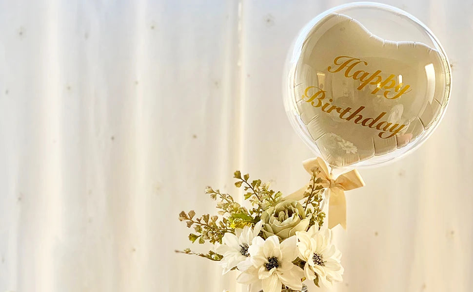 A clear bubble balloon shaped like a heart with gold lettering that reads Happy Birthday, decorated with white flowers.