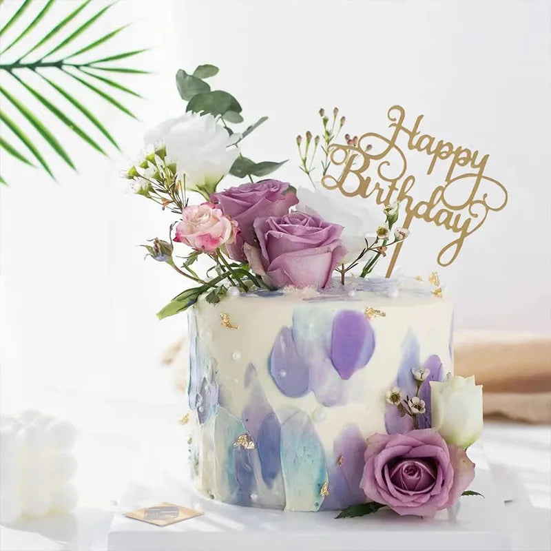 A white cake decorated with purple and blue watercolor frosting, adorned with pink roses and a gold birthday topper.