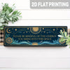 A rectangular dark blue metal tin sign featuring celestial designs and text on a white wooden surface.