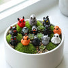 A collection of twelve mini resin Halloween duck figurines in various colors and textures arranged in a green moss setting.