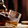 A hand holds a heavy base tumbler of amber whiskey on a wooden surface.