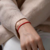A close-up of a hand wearing a handmade red string bracelet with a gold plated clasp, visible against a beige sweater sleeve.