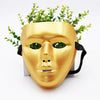 A golden masquerade mask with a face design and green foliage attached, displayed on a stand.