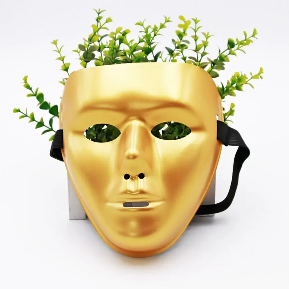 A golden masquerade mask with a face design and green foliage attached, displayed on a stand.