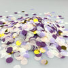 A pile of multicolor paper confetti consisting of purple, white, and gold pieces scattered on a light surface.