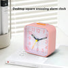 A pink desktop square snoozing alarm clock with a quartz movement resting on a white book.