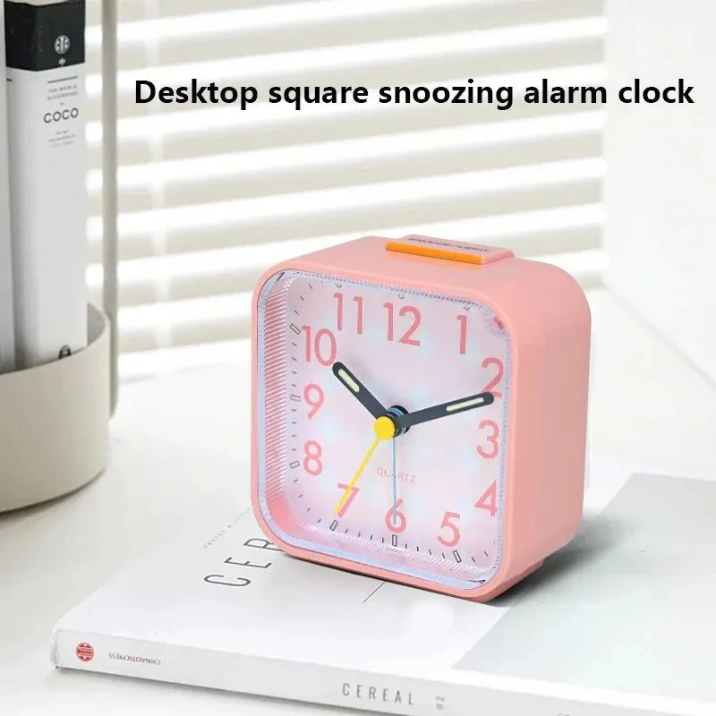 A pink desktop square snoozing alarm clock with a quartz movement resting on a white book.