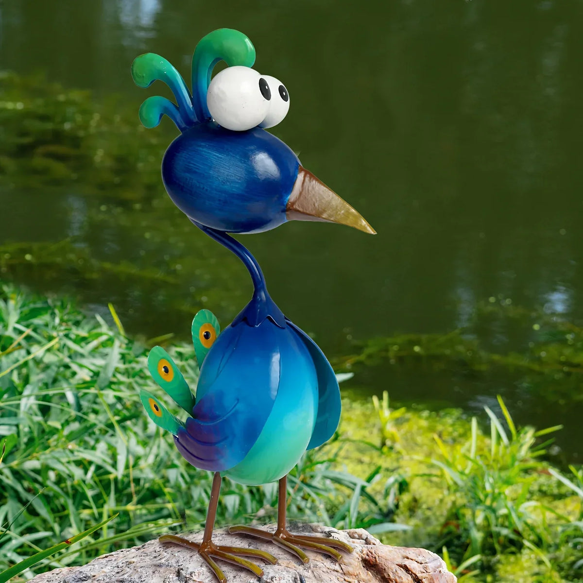 A blue metal peacock garden sculpture with a white head and green body standing on a rock near water.