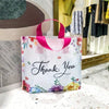 White floral thank you gift bags with pink handles displayed in a retail setting.