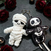 A white mummy skeleton plush keychain next to a dark gray one, set against a background of red roses.