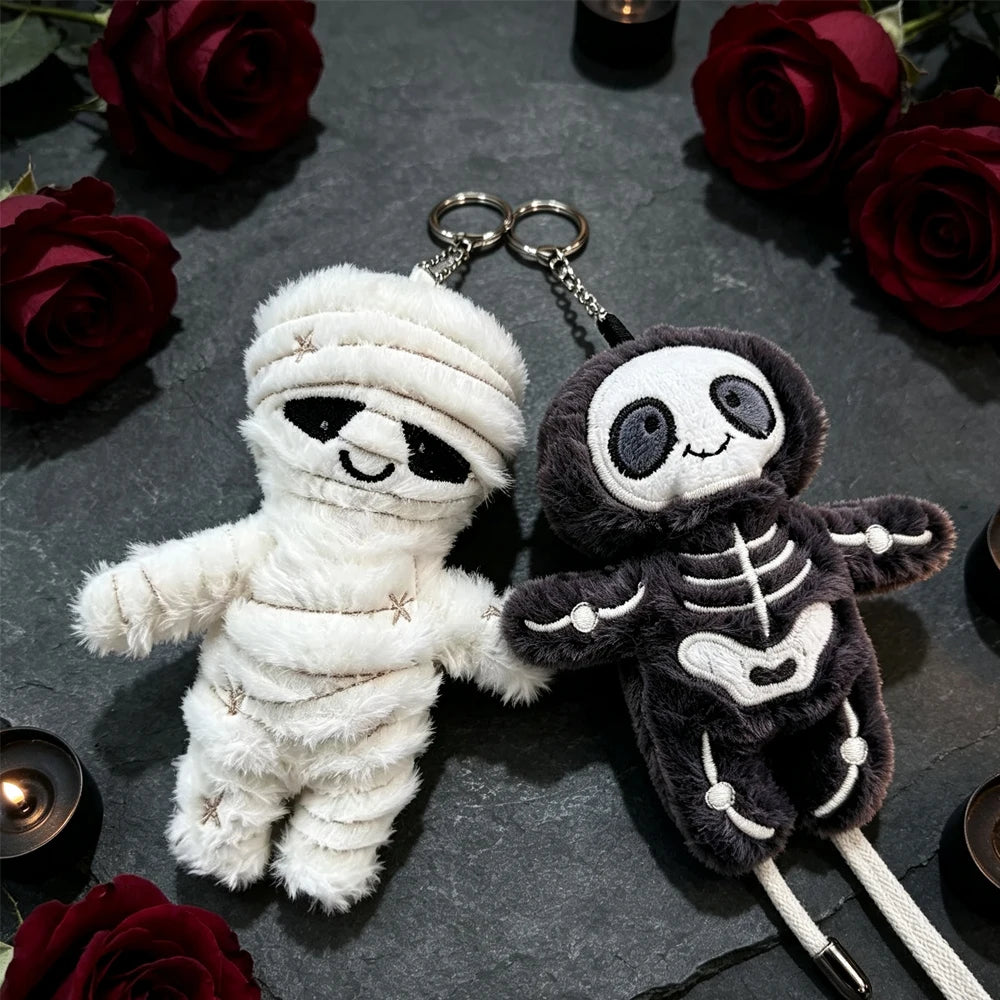 A white mummy skeleton plush keychain next to a dark gray one, set against a background of red roses.