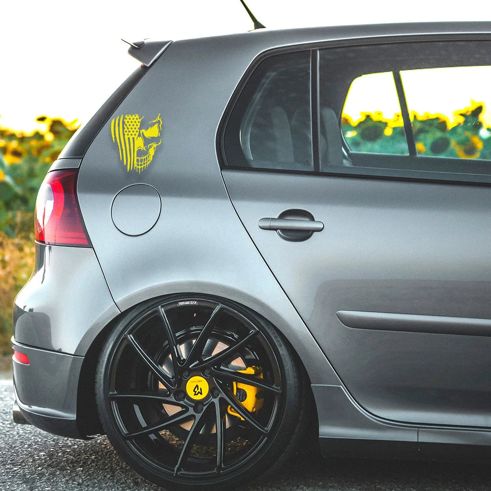 A gray car with a yellow patriotic skull decal on the rear fender and black aftermarket wheels.