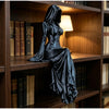 A black, flowing gown figurine of a hooded figure sitting on a wooden shelf among books.