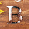 A silver metallic letter B cutout is placed on a wooden surface next to yellow flowers.