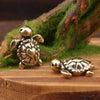 A handcrafted brass turtle figurine resting on a wooden surface next to mossy branches.