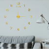 A modern wall clock with gold-colored numbers and hands is displayed on a light gray wall above a sofa.