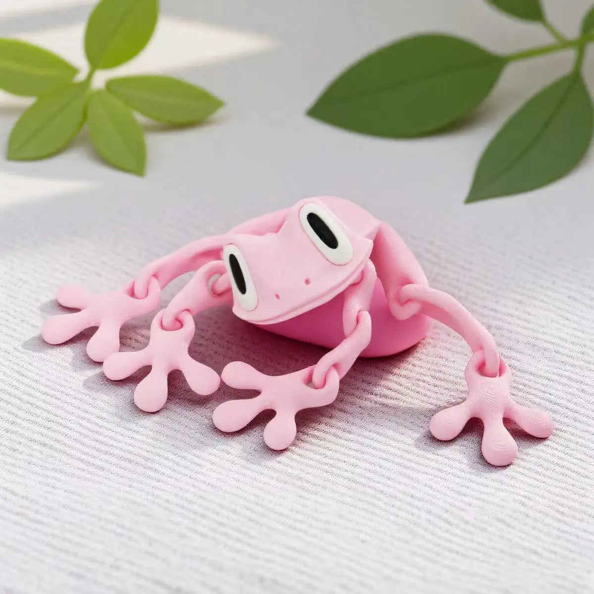 A pink, movable 3D printed tree frog ornament resting on a white textured surface with green leaves in the background.