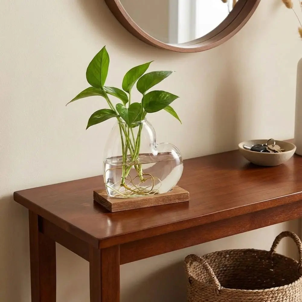 A green plant in a clear love-shaped glass vase sits on a wooden stand on a dark brown table.
