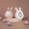 A pink mini LED cartoon alarm clock night light with rabbit ears sitting on a round stand.