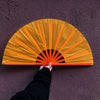 A hand is opening a yellow Tai Chi performance fan with orange ribs against a purple wall.