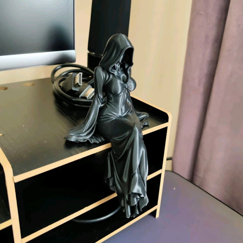 A black desktop figurine of a hooded female figure in a flowing gown, displayed on a dark wooden shelf.
